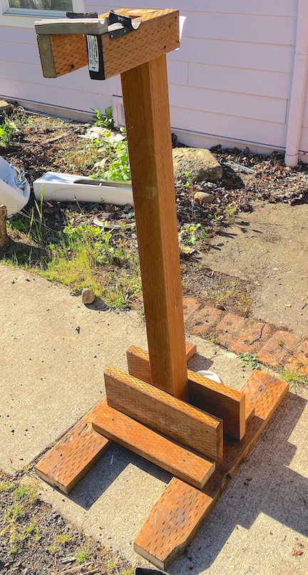 A bike repair stand made out of pressure treated wood. It has two flat 2x4 legs as a base, a single 4x4 as a center post, and two 2x4 arms coming out from the top for holding the bike up.