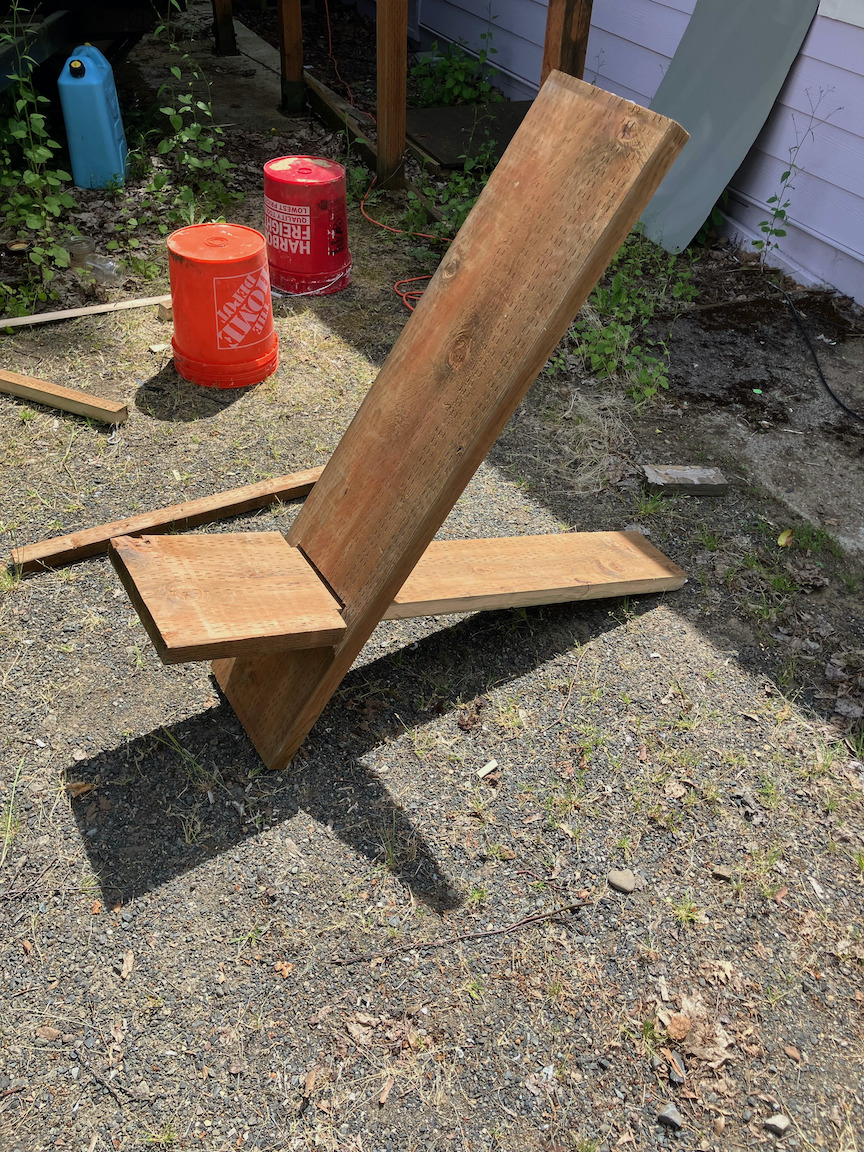 Outside resting on some gravel is this minimalist chair. It's an oblong 'X' shape, with one intersecting the other by going right through the center. On the more horizontal part, there is a small section coming out of the front at an upwards angle for the seat, with a long section going out behind. On the more vertical part, the back of the chair is tall and at a slight angle backwards.